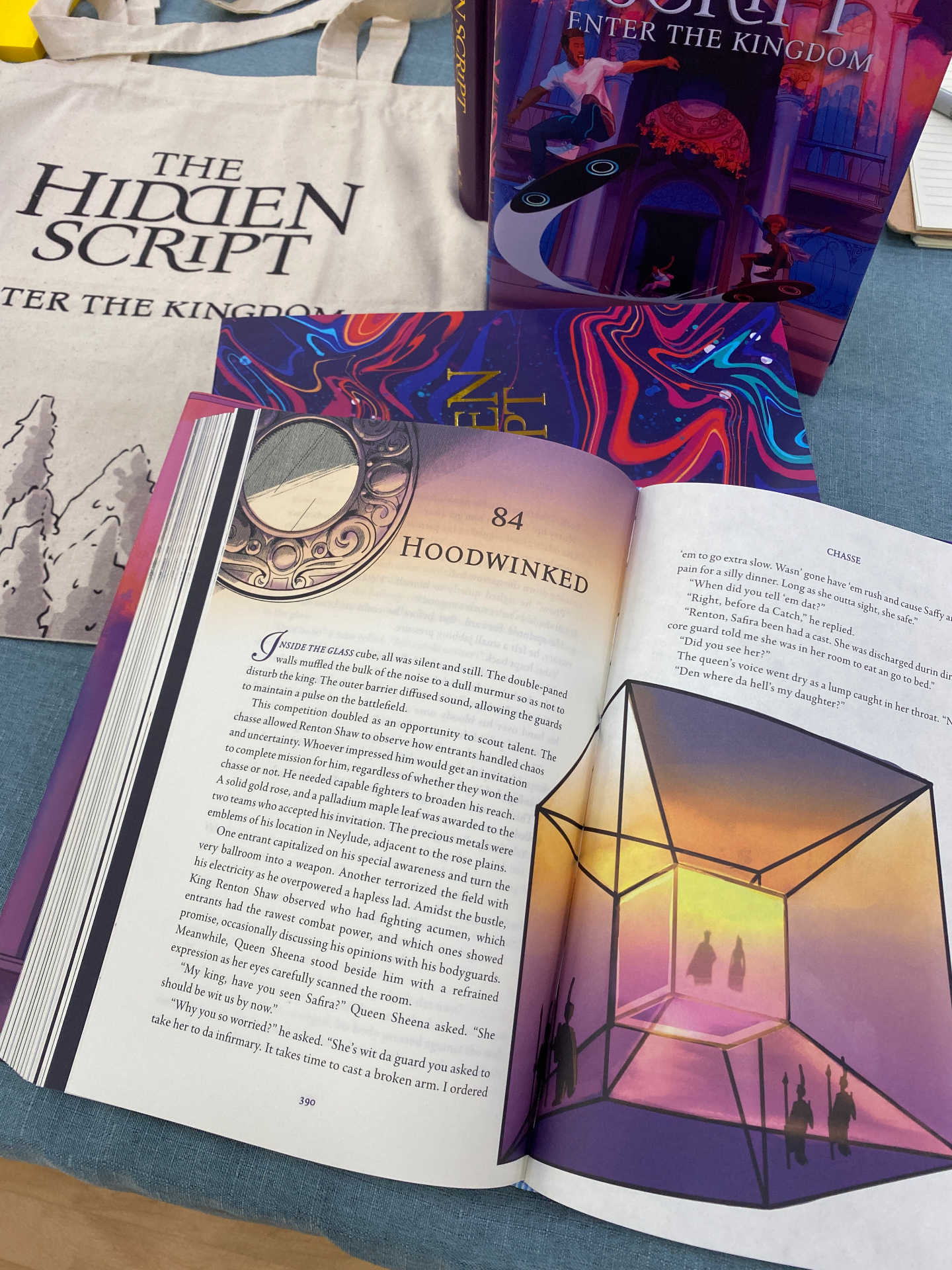 The Hidden Script book opened to chapter 84 Hoodwinked, with cover, tote bag, and convention props